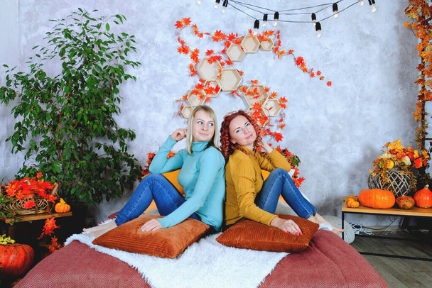 Two lovely young women, sisters, friends in warm sweaters are sitting in the bed at a cozy home, laughing, enjoying. Home interior with autumn decorations, orange leaves, flowers, pumpkins.  - Zdjęcie, obraz