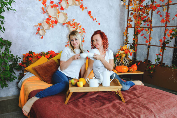 Дві прекрасні молоді жінки, сестри, друзі в теплих светрах сидять у ліжку в затишному будинку, сміються, насолоджуються. Інтер'єр будинку з осінніми прикрасами, апельсиновим листям, квітами, гарбузами
.  - Фото, зображення