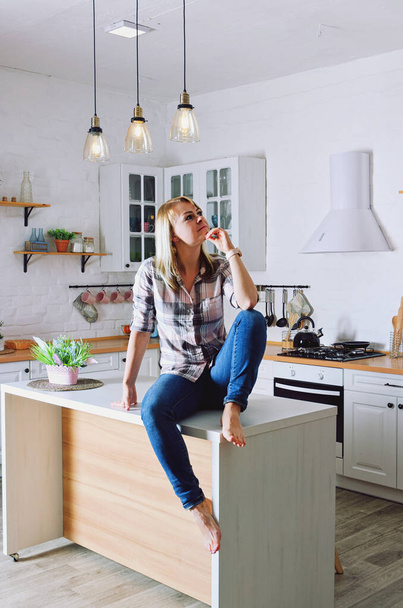 Mladá roztomilá blondýnka 30 + v modrých džínách a košili sedí a relaxuje ve své světlé čisté kuchyni. Zdravý šťastný životní styl. Volný čas. - Fotografie, Obrázek