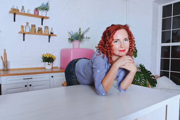 Fiatal aranyos vörös hajú göndör nő 30 + kék farmer és egy ing ül, és pihentető az ő világos tiszta konyha. Egészséges, boldog életmód. Szabadidő. Háziasszony.  - Fotó, kép