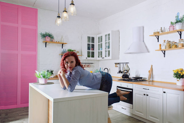 Young cute redhead curly woman 30+ in blue jeans and a shirt sitting and relaxing in her bright clean kitchen. Healthy happy lifestyle. Leisure. Housewife.  - Foto, Imagen