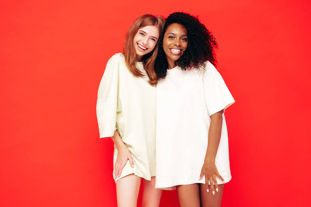 Two young beautiful smiling international hipster female in trendy summer clothes. Sexy carefree women posing near red wall in studio. Positive models having fun. Concept of friendship - Foto, immagini