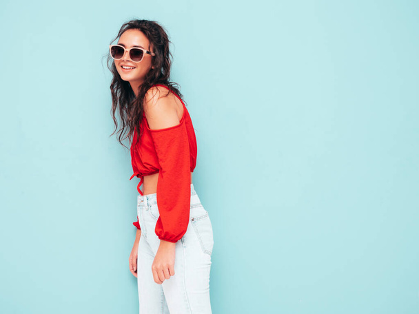 Young beautiful smiling female in trendy summer red top and jeans clothes. Sexy carefree woman posing near blue wall in studio. Positive brunette model having fun. Cheerful and happy. In sunglasses - Foto, imagen