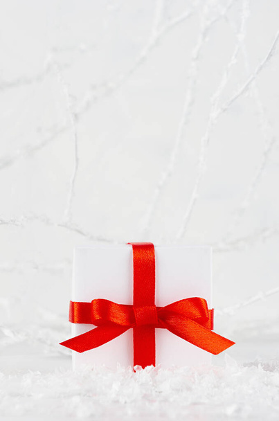 Festive christmas single gift box with red ribbon and bow in decorative snowdrift in fairy soft light winter forest with white frosty branches, copy space, vertical. Merry holiday New Year background. - Foto, imagen
