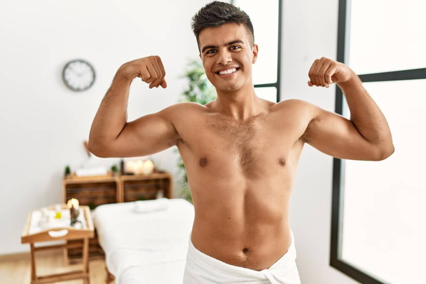 Young hispanic man standing shirtless at spa center showing arms muscles smiling proud. fitness concept.  - Photo, image