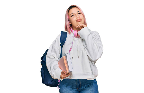 Latino nainen vaaleanpunaiset hiukset yllään opiskelija reppu ja kuulokkeet käsin leuka ajatella kysymys, mietteliäs ilme. Hän hymyili huomaavaisesti. epävarma käsite.  - Valokuva, kuva
