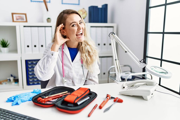 Young beautiful doctor woman with reflex hammer and medical instruments smiling with hand over ear listening an hearing to rumor or gossip. deafness concept.  - Photo, Image