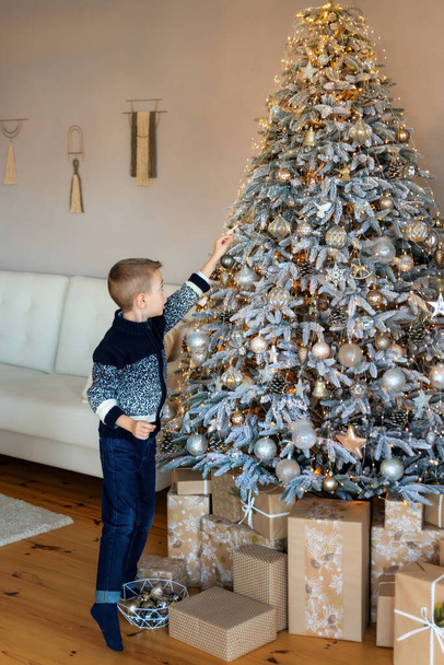 Little Caucasian boy in knitted sweaters and jeans decorate the Christmas tree. New Year or Christmas eve. Christmas tree decorated with New Year's toys. Vertical shot - 写真・画像