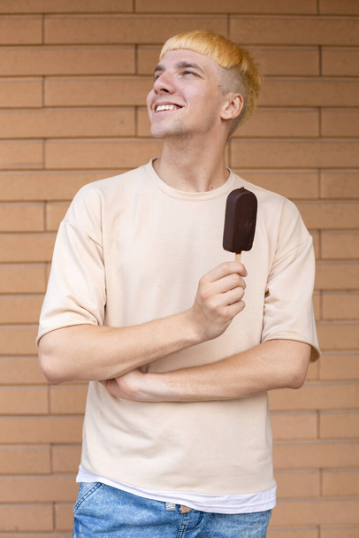 Summer, junk food, sweets and people concept - A happy Caucasian man dressed in a beige T-shirt and holding a chocolate ice cream on a stick looking away outdoors on a background of a brick wall  - Fotografie, Obrázek