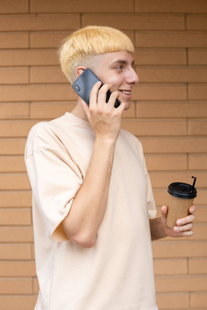 Estilo de vida, negócios, tecnologia e conceito de pessoas - Homem europeu sorridente vestido com uma camiseta bege, segurando uma xícara de café na mão e falando ao telefone ao ar livre em um fundo de parede de tijolo - Foto, Imagem