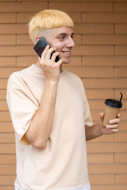 Lifestyle, business, technology, people concept - Een glimlachende Europese man gekleed in een beige T-shirt, met een kop koffie in de hand en praten aan de telefoon met collega op een bakstenen muur achtergrond - Foto, afbeelding