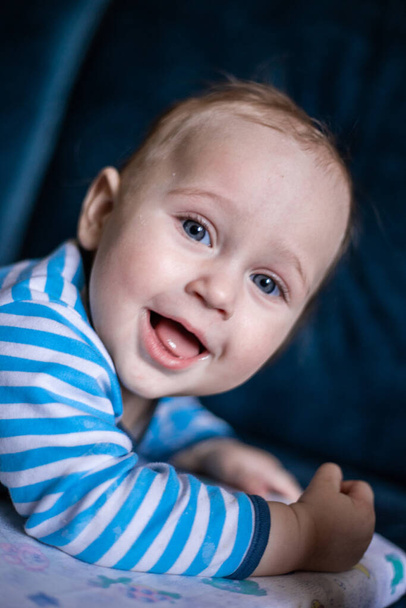 Un niño pequeño con los ojos azules en una blusa rayada yace sobre su estómago sobre un fondo borroso y sonríe mirando a la cámara. - Foto, imagen