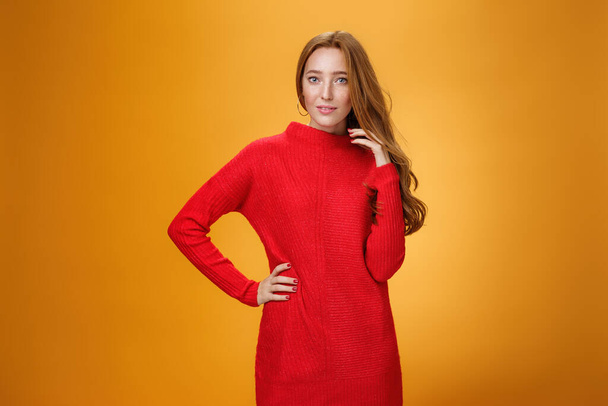 Atraktivní smyslné a romantické zrzka žena v červené pletené elegantní šaty drží ruku na boku s tajemným úsměvem jako flirtování hrát s vlasy zírá na kameru přes oranžové pozadí - Fotografie, Obrázek