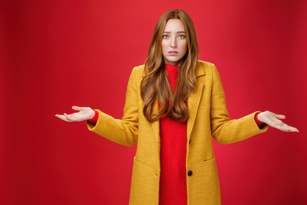 Upset and confused redhead girlfriend cannot understand what happened shrugging with spread hands sideways and clueless grimace, being unaware why, posing concerned over red wall - Photo, Image