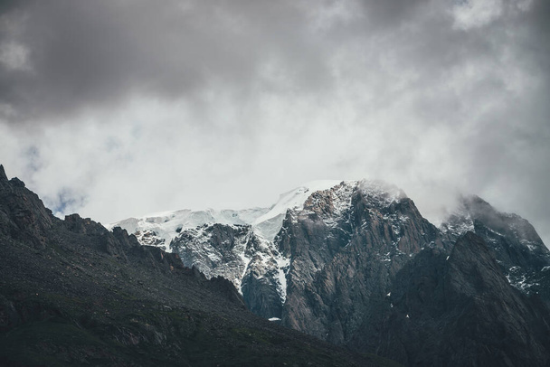 Dark atmospheric surreal landscape with dark rocky mountain top in low clouds in gray cloudy sky. Gray low cloud on high pinnacle. High black rock with snow in low clouds. Surrealist gloomy mountains. - Photo, Image