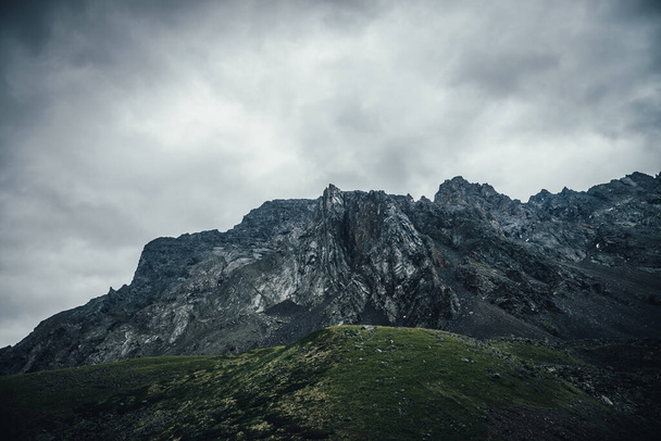 灰色の曇りの空の下で岩の山の壁と暗い大気の風景。鉛灰色の曇りの空のロッキー頂点。曇りの天気で暗い山のピーク。高い山と悲観的なミニマリストの風景 - 写真・画像