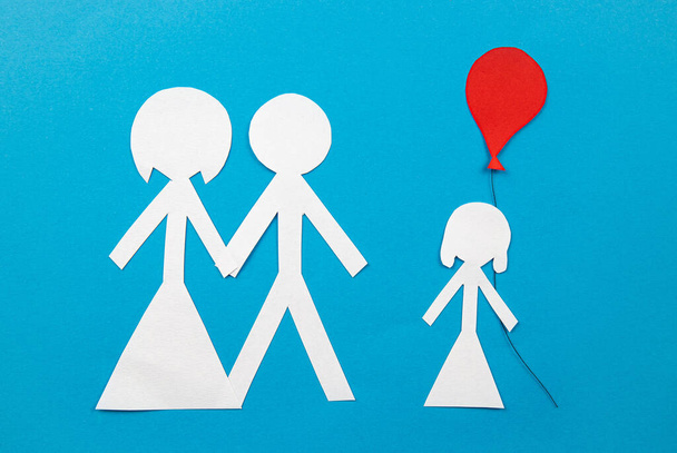 Conceptual image of adoption. Couple and child cut out of an white paper on blue background, indoors, studio shot. Child holding red balloon. Copy space. - Photo, Image