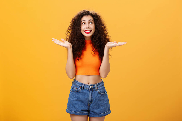 Silly and clueless carefree cute girl with curly hairstyle in cropped top and shorts shrugging with palms spread aside smiling unaware and gazing at upper right corner uncertain and uninvolved - Фото, изображение