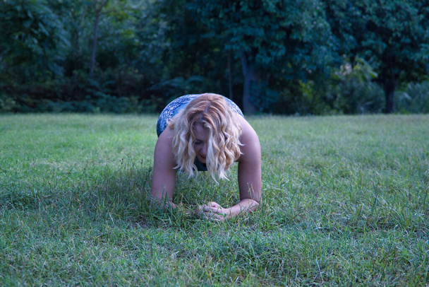 Молодая блондинка в спортивной одежде, делает брюшную доску, видна спереди посреди газона в парке. Концепция живота доска, фитнес, тренажерный зал, пышная девушка. - Фото, изображение