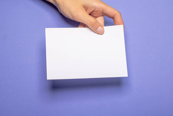 female hand holds a blank piece of paper with copy space in front of a violet colored background cardboard - Φωτογραφία, εικόνα