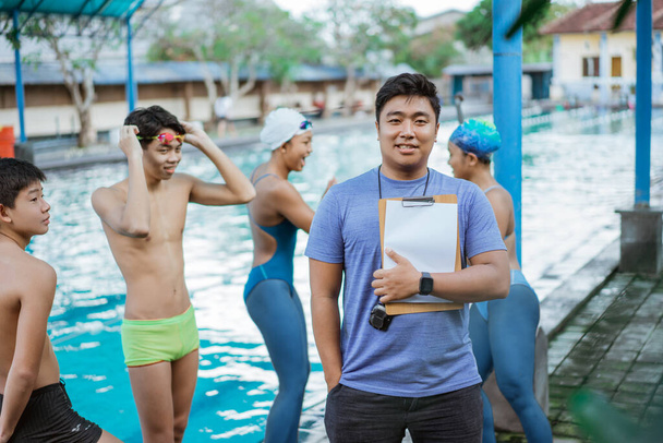 a swimming trainer smiling with one hand in pocket while holding a clipboard with his students - Valokuva, kuva