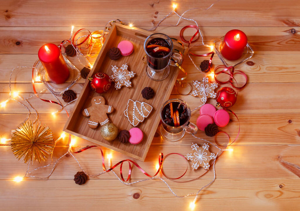 Dos copas de vino caliente, galletas de Navidad, velas, decoraciones, macarrones de fresa, galletas de chocolate y luces de Navidad brillantes en la mesa de madera. Vista superior.   - Foto, Imagen