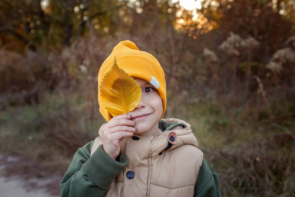 Vtipný chlapeček ve žlutém klobouku zakrývající jedno oko se žlutým listem během podzimní rodinné procházky - Fotografie, Obrázek