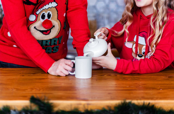 Isä ja tytär punaisissa joulupuseroissa. Tytär kaataa teetä teekannusta mukiin. Kuva: - Valokuva, kuva