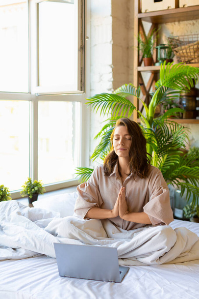 ラップトップモニターの前のベッドで瞑想する大人の女性の垂直写真。ヨガの練習. - 写真・画像