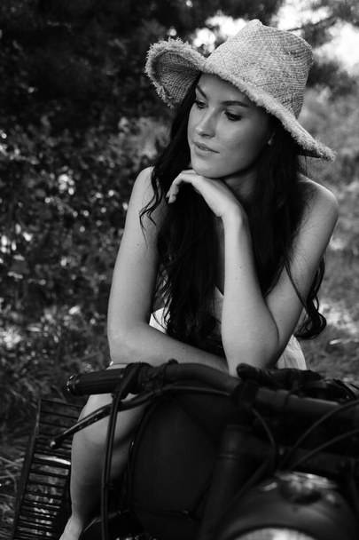 mladá sexy žena s dlouhými vlnitými vlasy v letních šatech a slaměný klobouk pózuje v blízkosti motocyklu. Černobílá fotografie - Fotografie, Obrázek