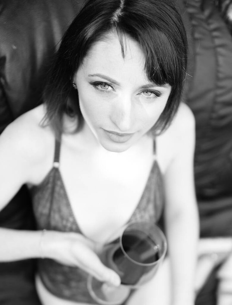 Mulher morena bonita jovem vestido em lingerie escura segurando vidro de vinho e posando em sofá de couro perto de casa de madeira ao ar livre  - Foto, Imagem