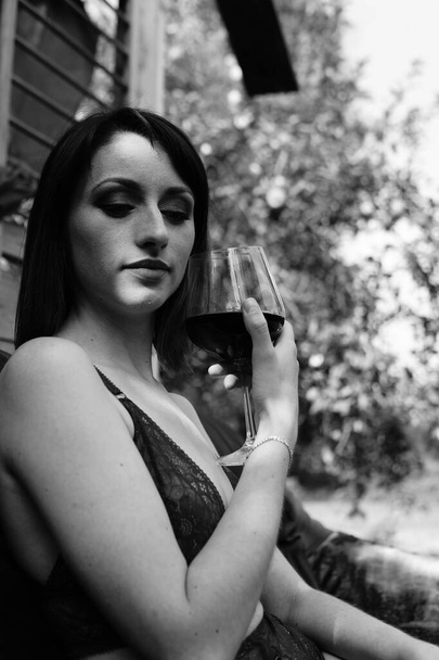 Csinos fiatal barna nő öltözött sötét fehérnemű kezében üveg bor és pózol a bőr kanapén közelében faház szabadban  - Fotó, kép