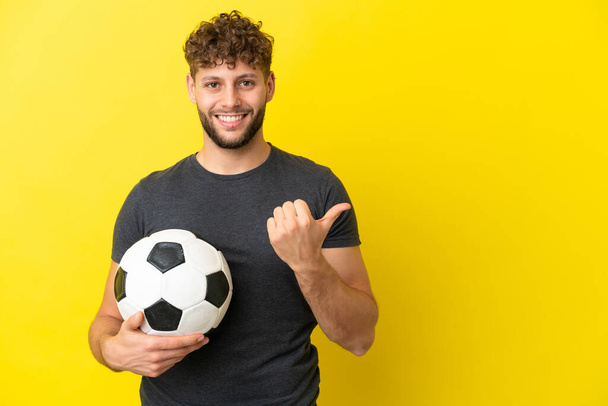 Όμορφος νεαρός ποδοσφαιριστής απομονωμένος σε κίτρινο φόντο που δείχνει προς τα πλάγια για να παρουσιάσει ένα προϊόν - Φωτογραφία, εικόνα