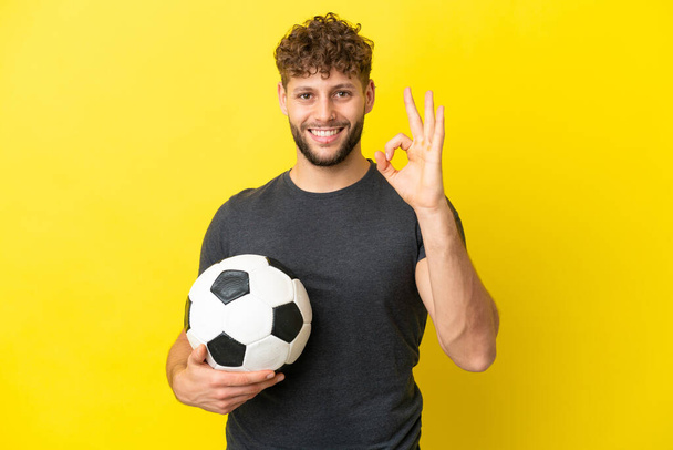Όμορφος νεαρός ποδοσφαιριστής άνδρας απομονωμένος σε κίτρινο φόντο δείχνει ok σημάδι με τα δάχτυλα - Φωτογραφία, εικόνα