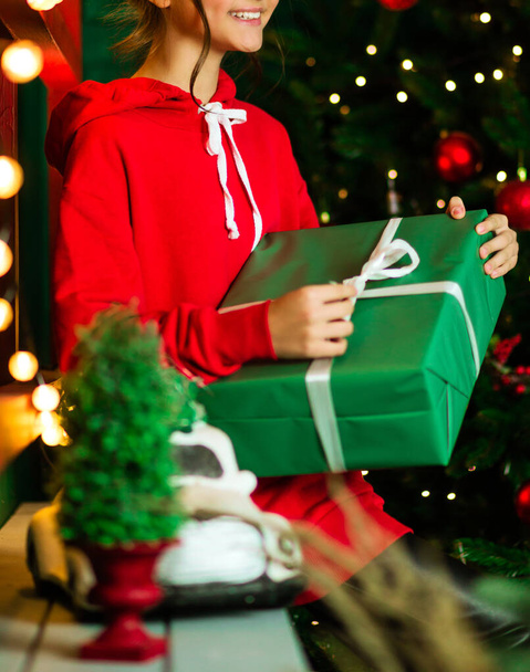 Une fille en vélo rouge s'assoit sur un canapé vert et ouvre un cadeau enveloppé dans du papier d'emballage vert. Photographie - Photo, image