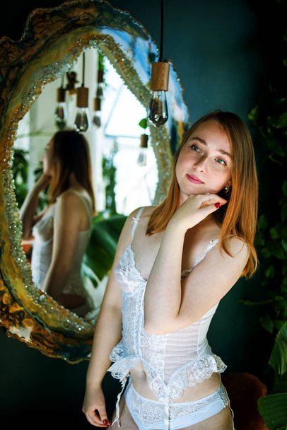 Красивая очаровательная молодая блондинка в белом сексуальном белье позирует в антикварном интерьере в роскошной квартире перед зеркалом - Фото, изображение