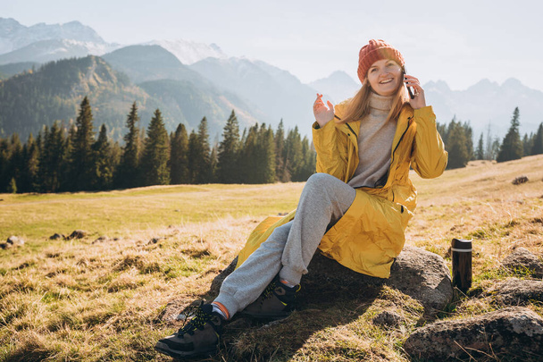 Jeune femme blonde assise et parlant par smartphone dans les montagnes. Liberté, bonheur, concept de voyage et vacances, activités de plein air - Photo, image