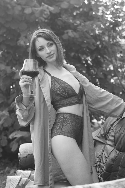 Όμορφη νεαρή μελαχρινή γυναίκα ντυμένη με σκούρα εσώρουχα κρατώντας ένα ποτήρι κρασί και ποζάροντας σε δερμάτινο καναπέ κοντά σε ξύλινο σπίτι εξωτερική  - Φωτογραφία, εικόνα