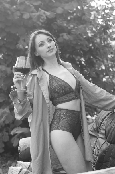 Bella giovane donna bruna vestita in lingerie scura che tiene un bicchiere di vino e posa sul divano in pelle vicino alla casa in legno all'aperto  - Foto, immagini