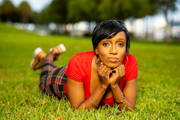 Foto de una hermosa modelo femenina posando al aire libre en una escena del parque. Acostado en la hierba con un top naranja y pantalones a cuadros - Foto, imagen