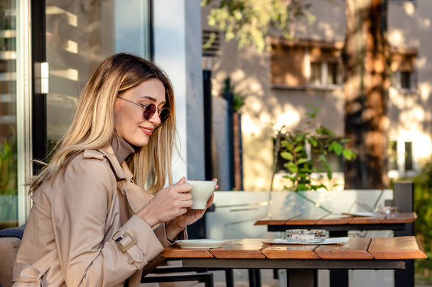Красивая блондинка пьет кофе в кафе на тротуаре, в солнечных очках в солнечный осенний день. - Фото, изображение