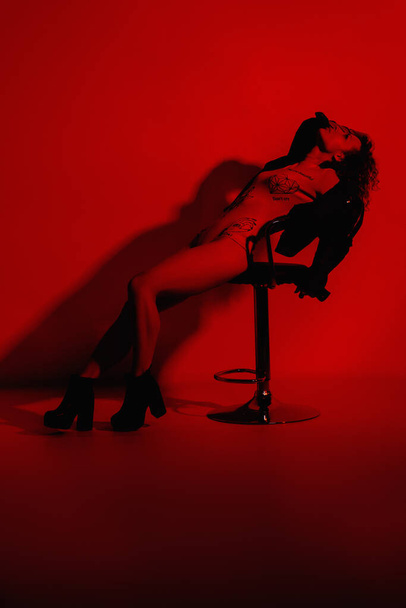 Modelka w stylowych ubraniach w neonach w studio. Studio portret seksownej kobiety w skórzanej kurtce, body suit i butach na czerwonym tle ultrafioletowym w klubie nocnym - Zdjęcie, obraz