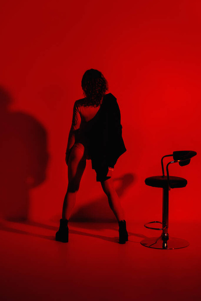 Divatmodell elegáns ruhákban a neon fények a stúdióban. Stúdió portré egy szexi nő bőrdzsekiben, testruha és csizma egy piros ultraibolya háttér egy nightclub - Fotó, kép