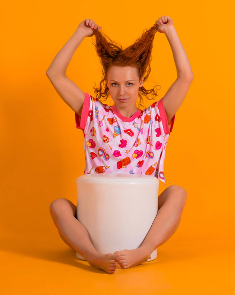 Φωτογραφία στούντιο μιας όμορφης κοκκινομάλλας με ροζ πιτζάμες σε κίτρινο φόντο. Μια γυναίκα κάθεται στην πολυθρόνα κρατώντας τα μαλλιά της και κοιτάζει την κάμερα.. - Φωτογραφία, εικόνα