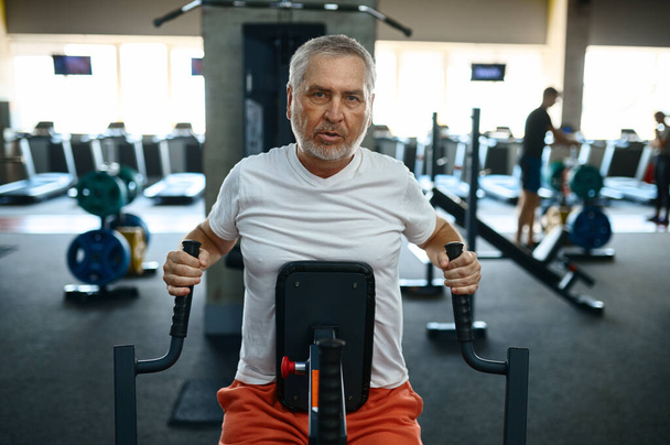 Starszy mężczyzna w odzieży sportowej na maszynie do ćwiczeń, wnętrze sali gimnastycznej na tle. Sportowy dziadek na treningu fitness w centrum sportowym. Zdrowy tryb życia, opieka zdrowotna - Zdjęcie, obraz