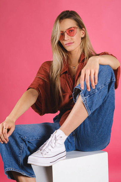 A vertical shot of an attractive Caucasian female wearing a red shirt and glasses against a pink wall - Foto, imagen