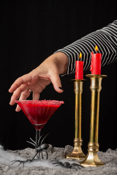 View of woman's hand on red cherry cocktail for Halloween with burning candles, on table with spiders, selective focus, black background, vertical - Photo, image