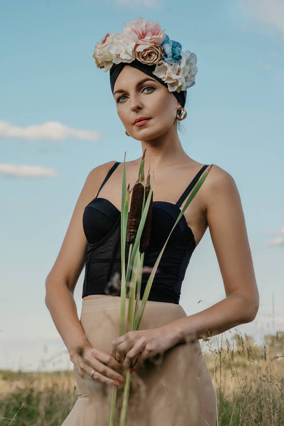 Girls in tops and shorts, in a hat and with a rim of flowers, posing against the background of nature. The first girl holds the stems of the reed. Another girl with a basket in which different flowers. - Φωτογραφία, εικόνα