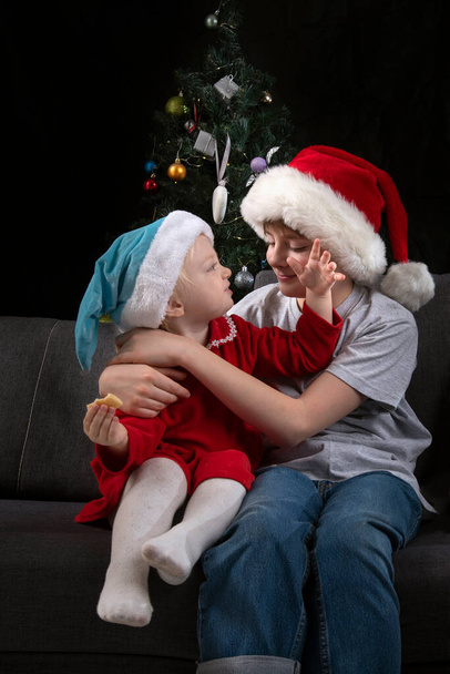 Αδελφός και αδελφή σε Σάντα καπέλα κάθονται στον καναπέ στο φόντο χριστουγεννιάτικο δέντρο. Κάθετο πλαίσιο. - Φωτογραφία, εικόνα