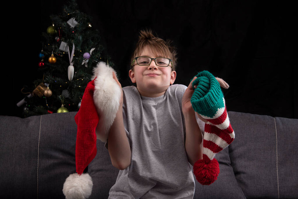 Boy is holding Santa hat and Christmas elf hat sitting on the sofa near the christmas tree. Role selection. - Photo, Image